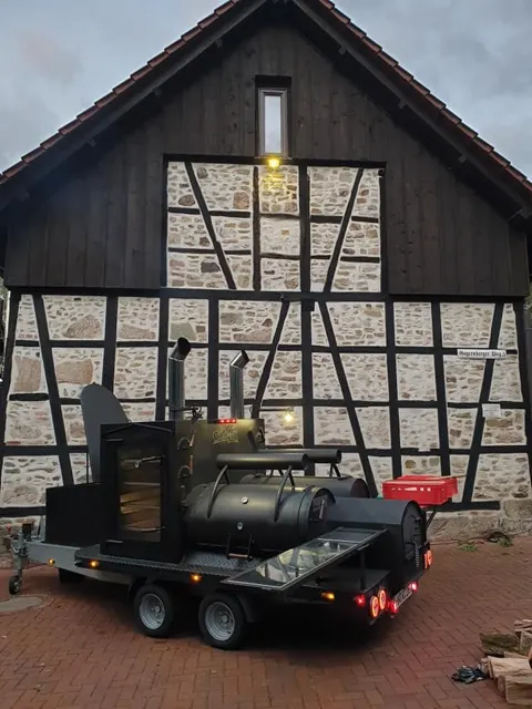 Emma, der XXL-Smoker von Fox BBQ: ein Anhänger mit großer liegender Räucherkammer, zwei Schornsteinen und Räucherschrank vor einer Fachwerkscheune.